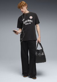 Sort bomulds T-shirt med grafik og teksten "Sankt Pauli", kombineret med vide sorte bukser. Holder en sort taske, har hovedtelefoner på og tjekker en telefon.