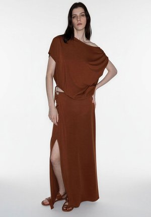 Mujer de pie con una mano en la cadera, vistiendo un top color óxido sin hombros y una falda larga a juego con una abertura hasta el muslo, y sandalias.