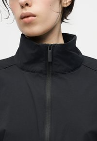 Κοντινό πλάνο ενός ατόμου που φοράει μαύρο μπουφάν με ψηλό γιακά, φερμουάρ κλειστό μέχρι το λαιμό, δείχνοντας το χερούλι του φερμουάρ με λογότυπο της μάρκας.