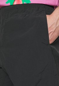 Calças leves pretas com uma textura suave, apresentando um bolso lateral e um cós elástico. A vista em close destaca os detalhes do tecido.