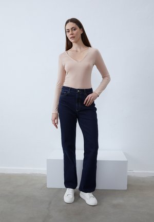 Μακρυμάνικη, ροζ V-neck μπλούζα συνδυασμένη με ψηλόμεσα σκούρα μπλε τζιν και λευκά sneakers, σε απλό, ανοιχτό φόντο.