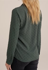 Grön långärmad skjorta med strukturerat, prickigt mönster. Har en krage, knappade ärmslut och en rundad nederkant. Tyget verkar lätt.