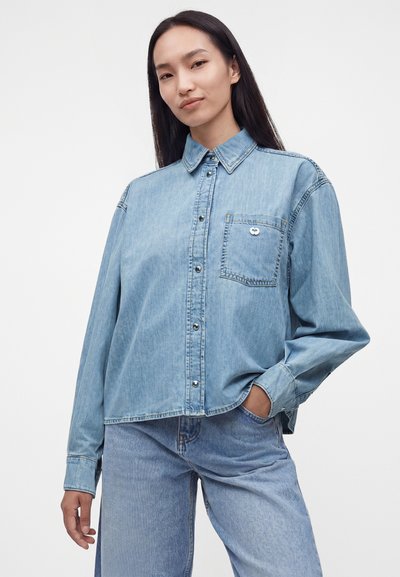 Jeune femme aux longs cheveux noirs portant une chemise en jean bleu clair ample et un jean assorti, une main dans la poche, fond neutre.