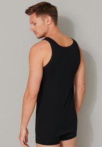 Zwarte katoen tanktop met brede bandjes, ronde halslijn en een aansluitende pasvorm. Het model draagt zwarte shorts en staat met de rug naar de camera.