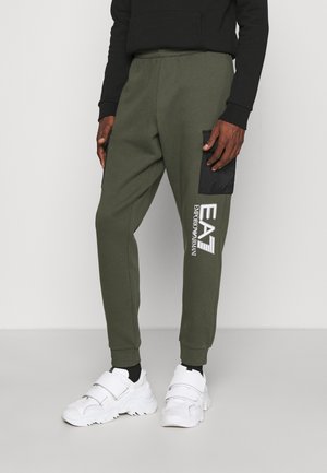 Man draagt olijfgroene joggingbroek met zwart zijvak en wit EA7 Emporio Armani-logo, gecombineerd met witte sneakers en zwarte sokken.