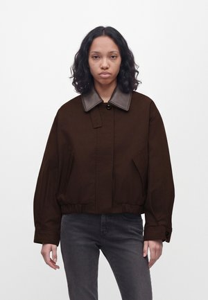Une femme porte une veste marron foncé à large col contrastant et un jean gris foncé sur un fond blanc uni.
