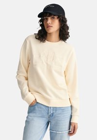 Cream-colored sweatshirt with embossed logo design, ribbed neckline and cuffs. Paired with light blue distressed jeans. Black cap with logo.