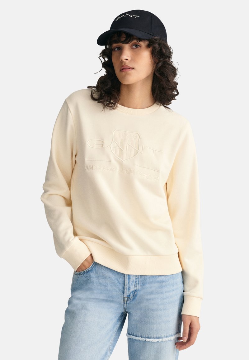 Cream-colored sweatshirt with embossed logo design, ribbed neckline and cuffs. Paired with light blue distressed jeans. Black cap with logo.