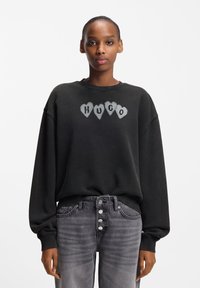 Juodas per dydį didelis džemperis su pilkomis širdies formos raidėmis "HUGO" ant krūtinės. Rankogaliai ir apatinė dalis su ribinėmis juostelėmis, derinami su pilkomis, išblukusiomis aukšto liemens džinsais.