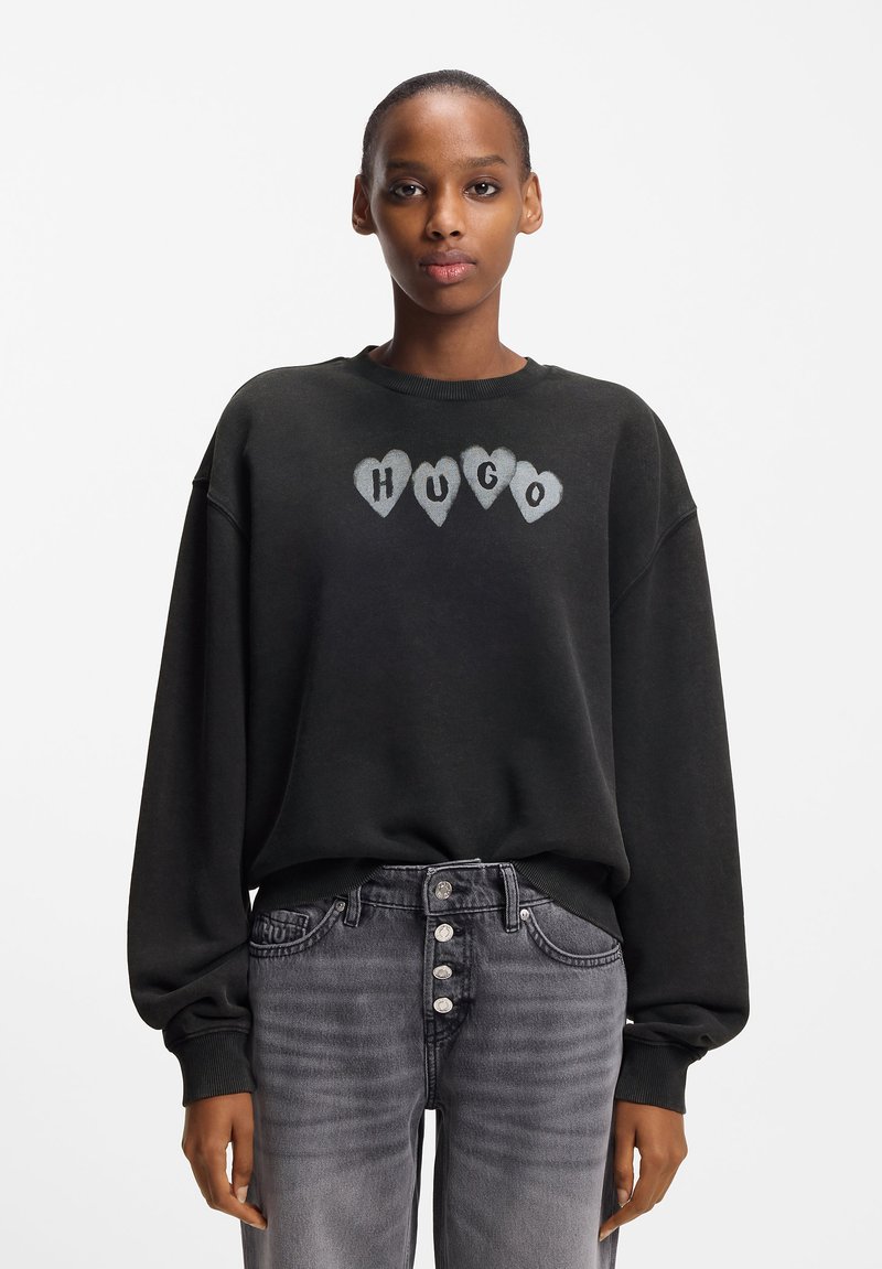 Juodas per dydį didelis džemperis su pilkomis širdies formos raidėmis "HUGO" ant krūtinės. Rankogaliai ir apatinė dalis su ribinėmis juostelėmis, derinami su pilkomis, išblukusiomis aukšto liemens džinsais.
