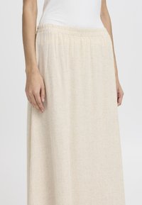 Jupe maxi beige clair en tissu tissé. Elle possède une taille élastique et une silhouette droite. Texture lisse avec des motifs minimaux.