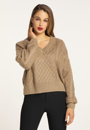 Donna con lunghi capelli castani indossa un maglione beige lavorato a trecce e pantaloni neri, posando con una mano vicino al collo su uno sfondo uniforme.