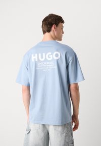 Ljusblå bomulls-t-shirt med kort ärm. Har stor vit text på baksidan: "HUGO," tillsammans med ytterligare tryckta detaljer nedanför.