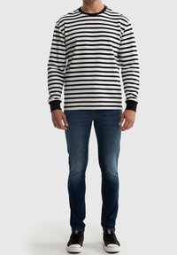 Hombre con camisa de rayas horizontales en blanco y negro de manga larga, jeans azules y zapatillas negras, de pie contra un fondo liso.