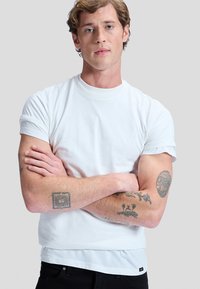 Young man with folded arms wearing a white t-shirt and black pants, showing tattoos on both forearms against a gray background.