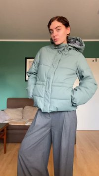 Puffet jakke i lysegrønn med hette, med glidelås foran og to sidelommer. Kombinert med løse grå bukser, mot en nøytral bakgrunn.