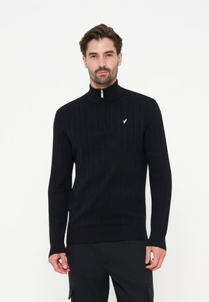 Mand iført en sort ribstrikket sweater med halv lynlås i kraven og et lille hvidt fuglelogo på brystet mod en ensfarvet hvid baggrund.