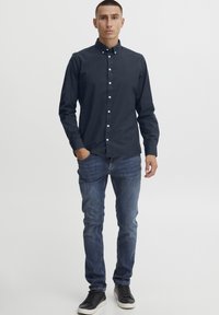 Temno modra srajca na gumb s ovratnikom, slim-fit kroj, dolgi rokavi; v kombinaciji z oprijetimi kavbojkami in črnimi supergami.