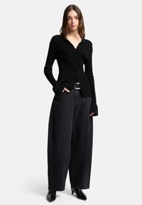 Zwart geribd gebreid vest met knoopsluiting aan de voorkant en lange mouwen. Gecombineerd met hooggetailleerde, wijd uitlopende zwarte jeans en puntige zwarte schoenen.