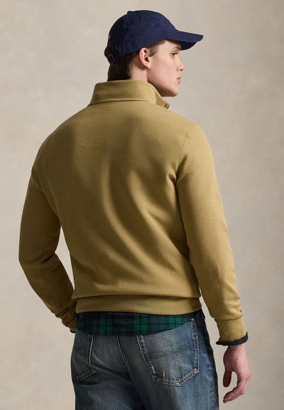 Vyras vilki tamsiai mėlynę kepurę, šviesiai rudą striukę ant žalios languotos marškinės ir mėlynas džinsas, žiūri į šoną prieš paprastą smėlio spalvos foną.