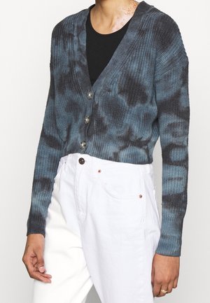 Osoba ubrana w niebiesko-czarny krótkim kardigan z wzorem tie-dye i guzikami, czarny top pod spodem oraz białe spodnie z wysokim stanem.