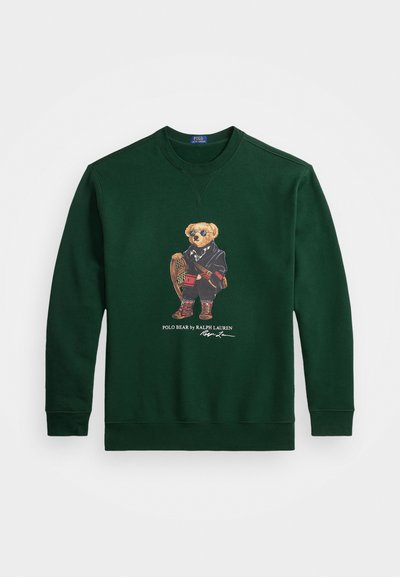 Zaļš džemperis ar lāča grafiku, kas valkā saulesbrilles un melnu apvalku, ar sniega kurpēm un zābakiem, kā arī tekstu "Polo Bear by Ralph Lauren".