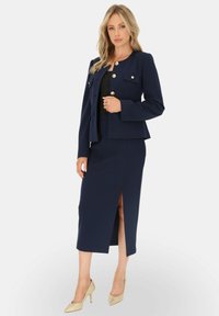 Granatowa marynarka z złotymi guzikami, połączona z dopasowaną spódnicą do kolan z rozcięciem. Całość dopełniają eleganckie buty.