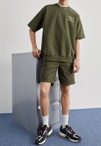 Sudadera verde oliva con un gran gráfico, combinada con shorts de cargo verdes oliva. El modelo lleva calcetas blancas y zapatillas multicolores, de pie junto a un archivador.
