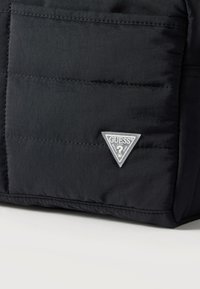 Γωνία από μαύρο υφαντό τσαντάκι με ραμμένες ραφές και μικρό τριγωνικό λογότυπο Guess U.S.A. σε λευκό και γκρι.