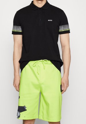 Mann trägt ein schwarzes Polohemd mit "BOSS"-Logo auf der Brust und gemusterten Ärmeln, kombiniert mit hellen limettengrünen Shorts mit Kordelzug.