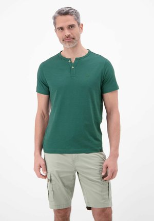 Mann mit grauen Haaren, der ein grünes Henley-Shirt mit kurzen Ärmeln und hellfarbige Cargo-Shorts trägt, steht vor einem schlichten weißen Hintergrund.