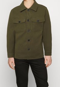 Olivengrøn jakke med knaplukning, to brystlommer og krave, lavet af et tekstureret stof.
