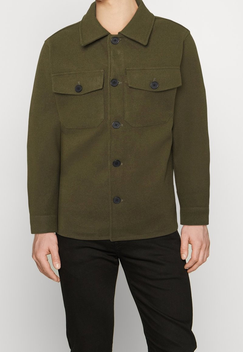 Olivengrøn jakke med knaplukning, to brystlommer og krave, lavet af et tekstureret stof.