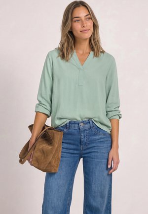 Une femme porte un chemisier vert pâle et un jean bleu, tenant un sac fourre-tout en daim marron sur un fond clair de studio.