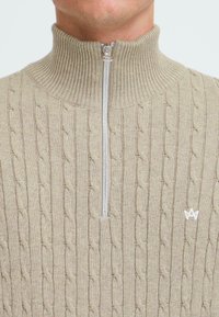 Maglione beige a trecce con colletto alto a costine e zip argentata. Presenta un piccolo logo bianco vicino al fondo destro.