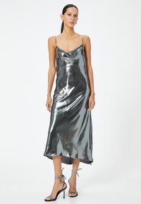 Metallisch graues Satin-Midikleid mit Spaghettiträgern, Cowl-Ausschnitt und einem leicht asymmetrischen Saum. Kombiniert mit schwarzen Riemchen-Absätzen.