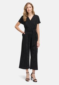 Combinaison noire avec un design à enroulement, manches courtes, détail de taille nouée et pantalons larges ; le tissu a un subtil éclat métallique.