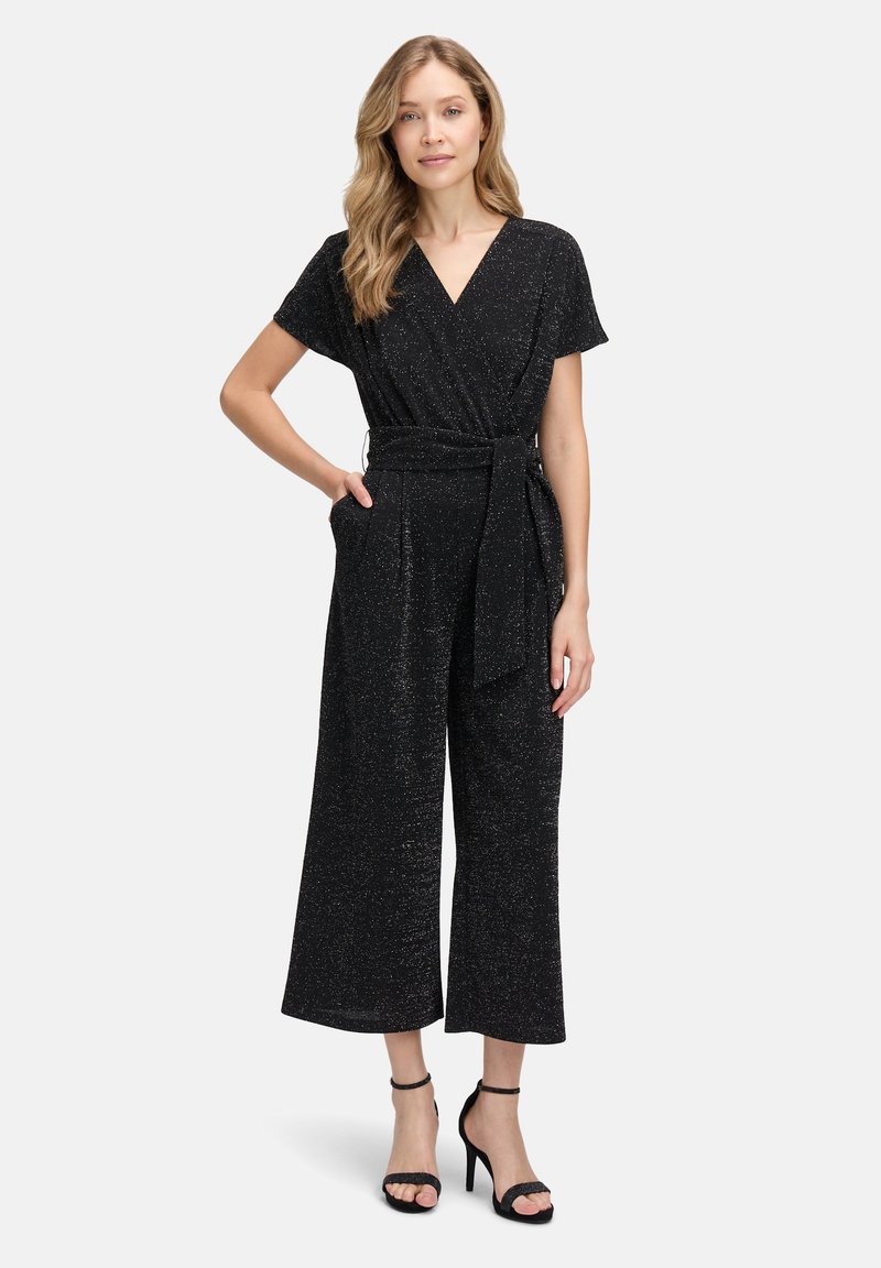 Combinaison noire avec un design à enroulement, manches courtes, détail de taille nouée et pantalons larges ; le tissu a un subtil éclat métallique.