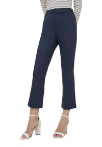 Pantaloni tailored blu navy con una linea dritta, caratterizzati da una texture liscia e un sottile lucido. Abbinati a sandali con tacco bianchi.