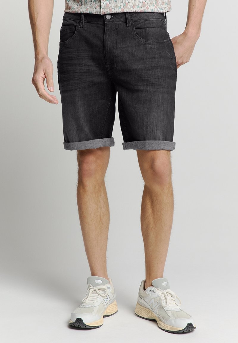Schwarze Denim-Shorts mit umgeschlagenen Säumen, mit frontalen Taschen und einem Knopfverschluss. Getragen mit hellen Sportschuhen.