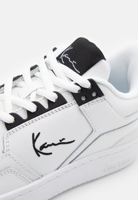 White leather sneaker with black accents, a padded tongue, and white laces. Features a signature in black on the side. Textured interior.