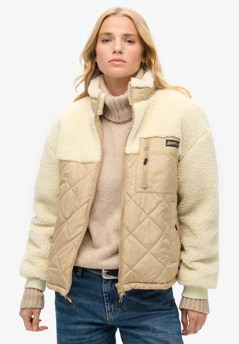 Veste matelassée beige avec des manches en fausse fourrure crème, poche poitrine zippée et poignets côtelés. Portée par-dessus un pull beige clair.