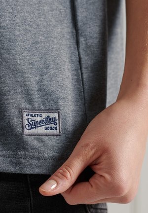 Grå bomulds T-shirt med et syet mærke med "ATHLETIC SUPERDRY GOODS." Blød tekstur, korte ærmer og en afslappet pasform.
