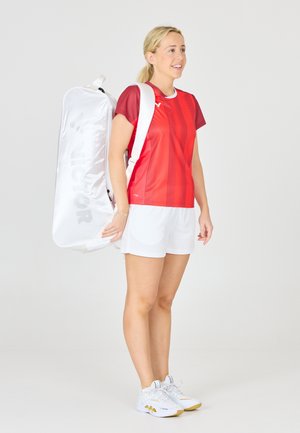 Rød sportsjersey med korte ærmer og hvide detaljer, hvide shorts og en hvid rygsæk med et logo. Model iført grå træningssko.