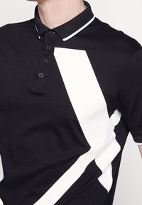 Homme portant un polo noir avec des rayures diagonales blanches et des boutons noirs, cadré en dessous du cou et au-dessus de la taille.