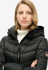 Zwarte pufferjack met rits en capuchon. Kenmerkt zich door de gestikte textuur en het Superdry-logo op de mouw. Gemaakt over een witte turtleneck.
