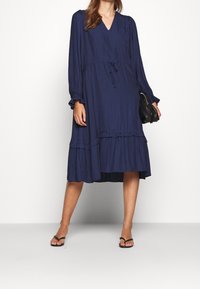 Navy modrošedé, dlouhé šaty s výstřihem do V, stahovací pasem a patrovým volánovým lemem. Vyrobené z texturované látky s vertikálními pruhy.