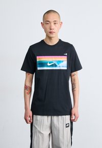 Svart bomull T-skjorte med et rektangulært flerfarget grafikk som har en blå bølge og hvit Nike-logo på foran. Rund hals design.