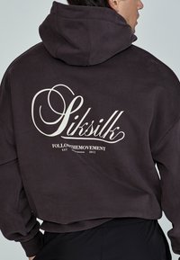 Hanorac gri închis cu un logo mare alb care prezintă cuvântul "Siksilk" și "FOLLOWTHEMOVEMENT" pe spate. Material moale, casual.