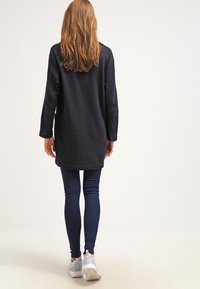 Tmavě modrý dlouhý fleece kabát, sahající do poloviny stehna, v kombinaci s námořnickými legínami a světlými teniskami. Hladká textura a volný střih.